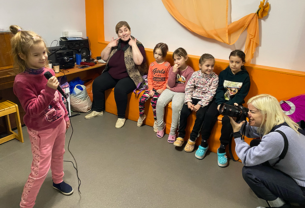 Four children and one adult woman sit in a row on a bench along an interior wall. A fifth child stands in front of a microphone, appearing to speak or perform, while a woman with a camera crouches in the foreground, possibly capturing the event. 