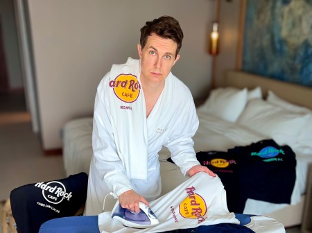 Matt Houchin, wearing a white bathrobe, stands at a table covered in Hard Rock Café T-shirts, ironing one of the shirts.