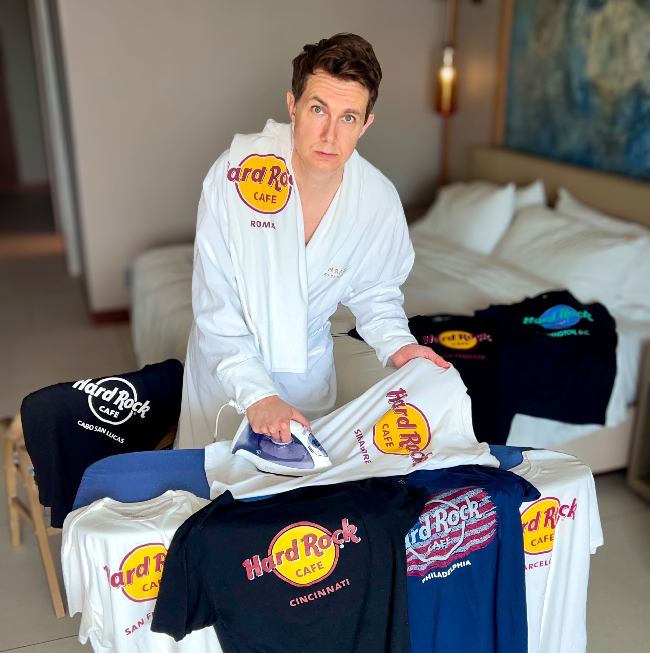 Matt Houchin, wearing a white bathrobe, stands at a table covered in Hard Rock Café T-shirts, ironing one of the shirts.