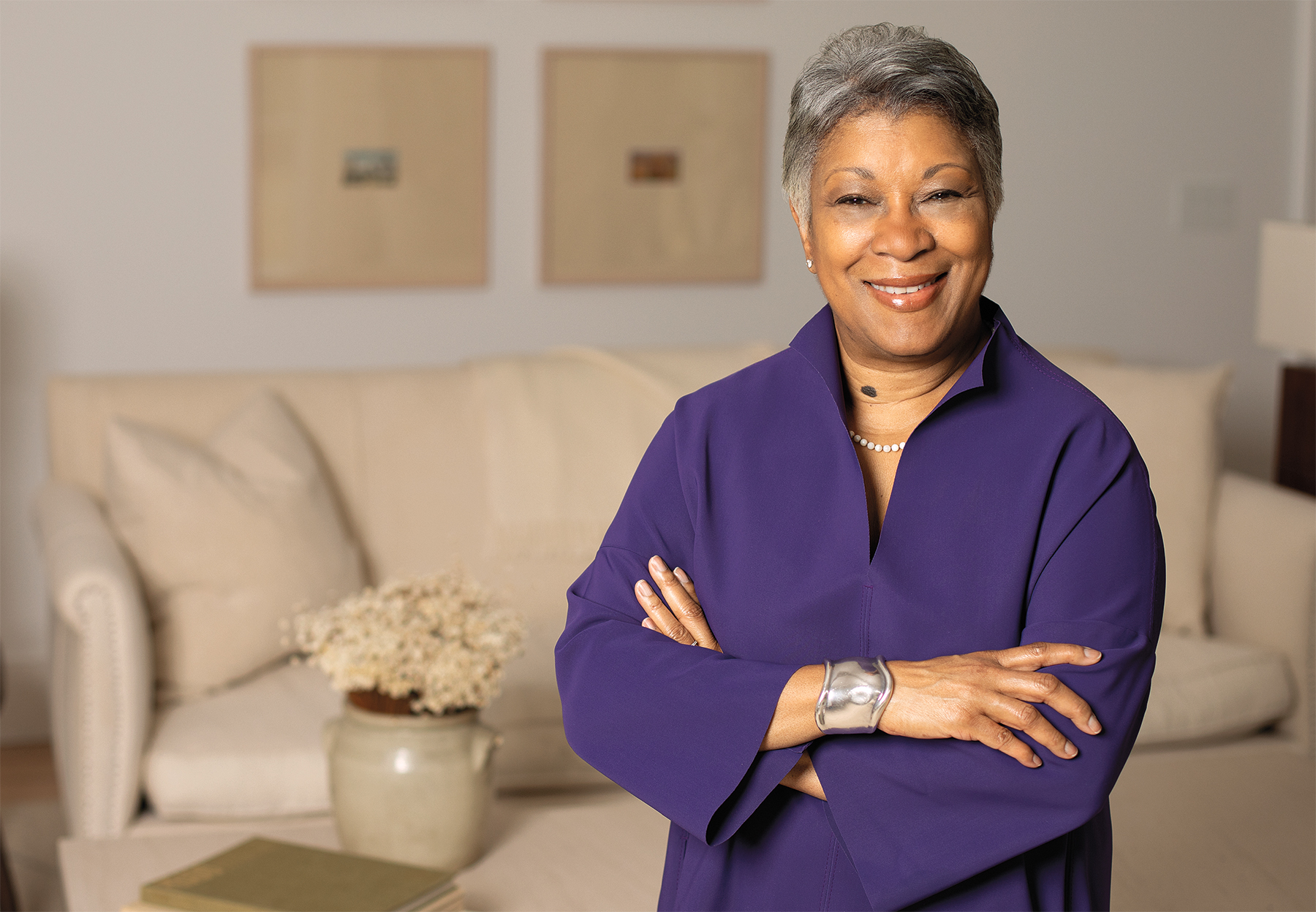 Renetta McCann stands with her arms folded, wearing a purple pantsuit and smiling at the camera. Behind her is a white couch and coffee table with books and flowers.