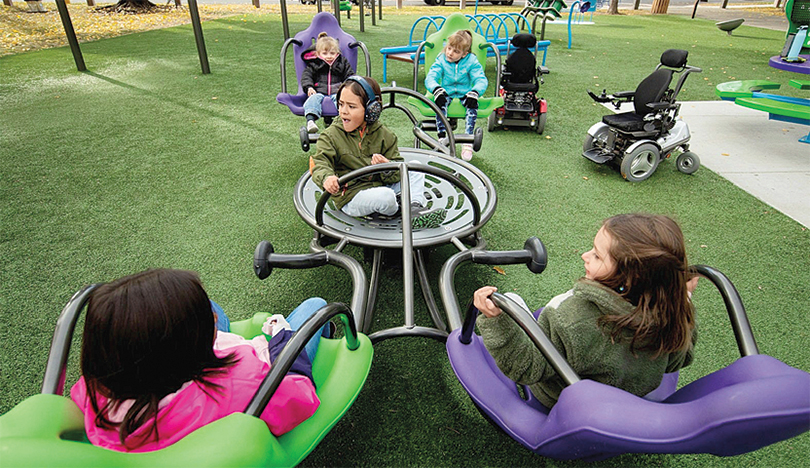 In a playground, five children sit on a see-saw that has four high back seats as well as a center saucer. In the background are two empty wheelchairs, which belong to a couple of the children. 