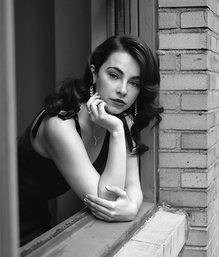 A black-and-white photo of Stella Cole leaning out of an apartment window in New York City. She is resting her chin on her right arm and her gaze looks away from the camera to her right. She is wearing a dress and a necklace.