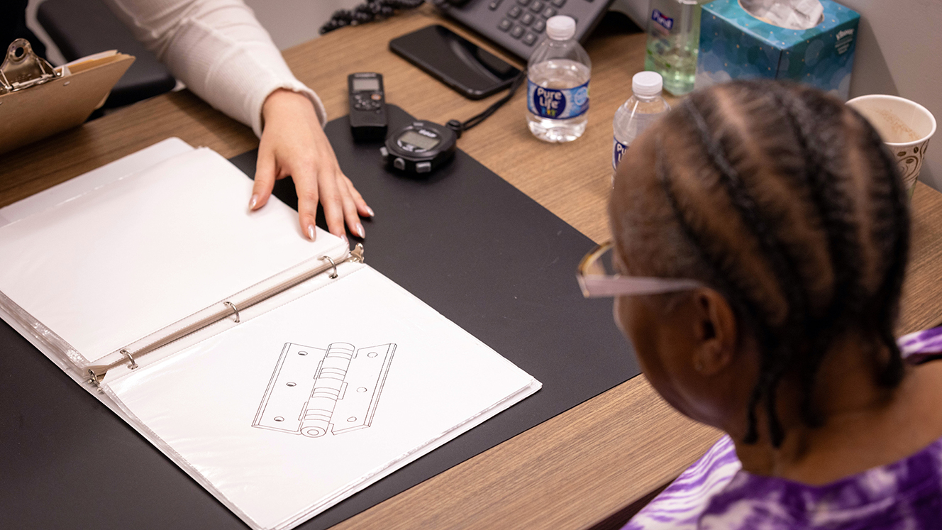 Yvonne Gregwor, wearing a purple tie-dyed t-shirt, looks down at a page in a binder as a researcher holds the binder open.