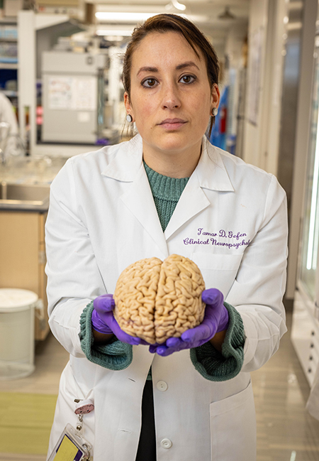 Tamar Gefen stands in a science lab, holding a brain in both hands and looking into the camera. She is wearing a white lab coat with a green shirt underneath and purple gloves.