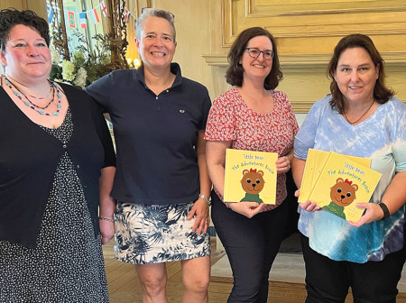 Katy Wills, Sarah Wills Carlsson, Jennifer Royall Anderson, Jennifer Burke and Valerie Banks Amster pose in front of a fireplace, smiling with their arms around each other while Anderson and Burke hold copies of ’ittle Bear: The Adventures Begin. 