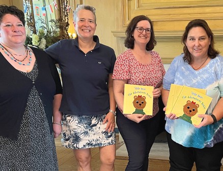 Katy Wills, Sarah Wills Carlsson, Jennifer Royall Anderson, Jennifer Burke and Valerie Banks Amster pose in front of a fireplace, smiling with their arms around each other while Anderson and Burke hold copies of ’ittle Bear: The Adventures Begin. 