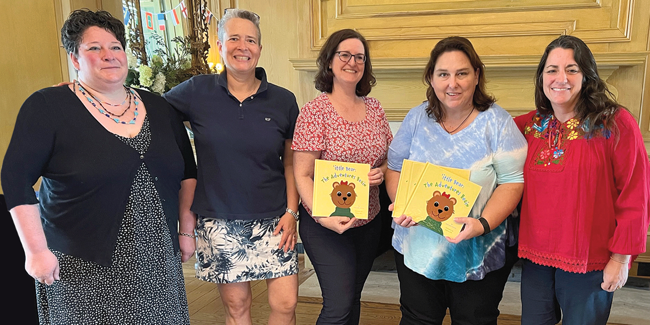 Katy Wills, Sarah Wills Carlsson, Jennifer Royall Anderson, Jennifer Burke and Valerie Banks Amster pose in front of a fireplace, smiling with their arms around each other while Anderson and Burke hold copies of ’ittle Bear: The Adventures Begin. 