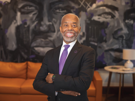 Will Evans stands with his arms folded, wearing a black suit and purple tie and smiling at the camera, with a brown couch and side table in the background.