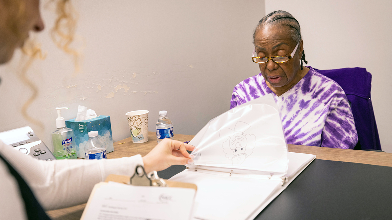 Yvonne Gregwor, wearing a purple tie-dyed t-shirt, sits at a table across from a researcher, looking down at a binder. The researcher takes notes on a clipboard with her right hand while flipping the page in the binder with her left hand, showing an image of a clown on one of the pages.