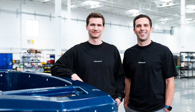 Ryan Cook, left, and Mitch Lee stand in a manufacturing facility. They both wear black T-shirts shirts that say “100% electric.” There are shelves in the background.  