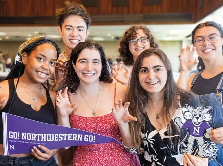 A group of six Northwestern seniors smile at the camera. Each of them is curling one of their hands into a Wildcat “claw.” The student on the far left holds a purple pennant with the words “Go! Northwestern Go! Homecoming and Reunion 2023” on it, followed by the school’s academic N. Two other students on the far right hold a cut-out image of Willie the Wildcat, who is wearing a purple shirt with the academic N on it. 