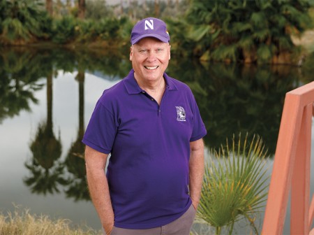 Marc McClellan stands near a body of water in California. He wears a purple polo shirt with Northwestern’s athletic logo on his upper left chest. He also wears a purple baseball cap displaying Northwestern’s athletic N. The opposite side of the water is lined with palm trees and lush greenery, which are reflected in the water’s surface. To McClellan’s left, part of a pedestrian bridge is visible. 