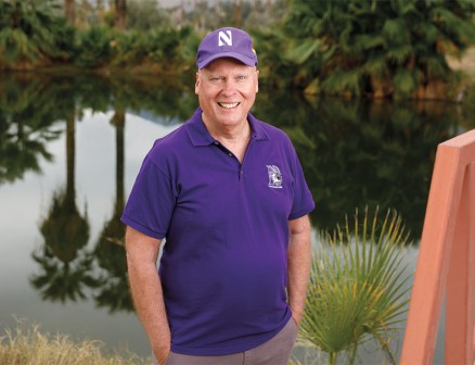 Marc McClellan stands near a body of water in California. He wears a purple polo shirt with Northwestern’s athletic logo on his upper left chest. He also wears a purple baseball cap displaying Northwestern’s athletic N. The opposite side of the water is lined with palm trees and lush greenery, which are reflected in the water’s surface. To McClellan’s left, part of a pedestrian bridge is visible. 