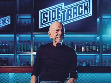 Art Johnston sits on a barstool in Sidetrack. He is smiling and looking slightly away from the camera to his left. Behind him is a neon sign displaying the name Sidetrack.