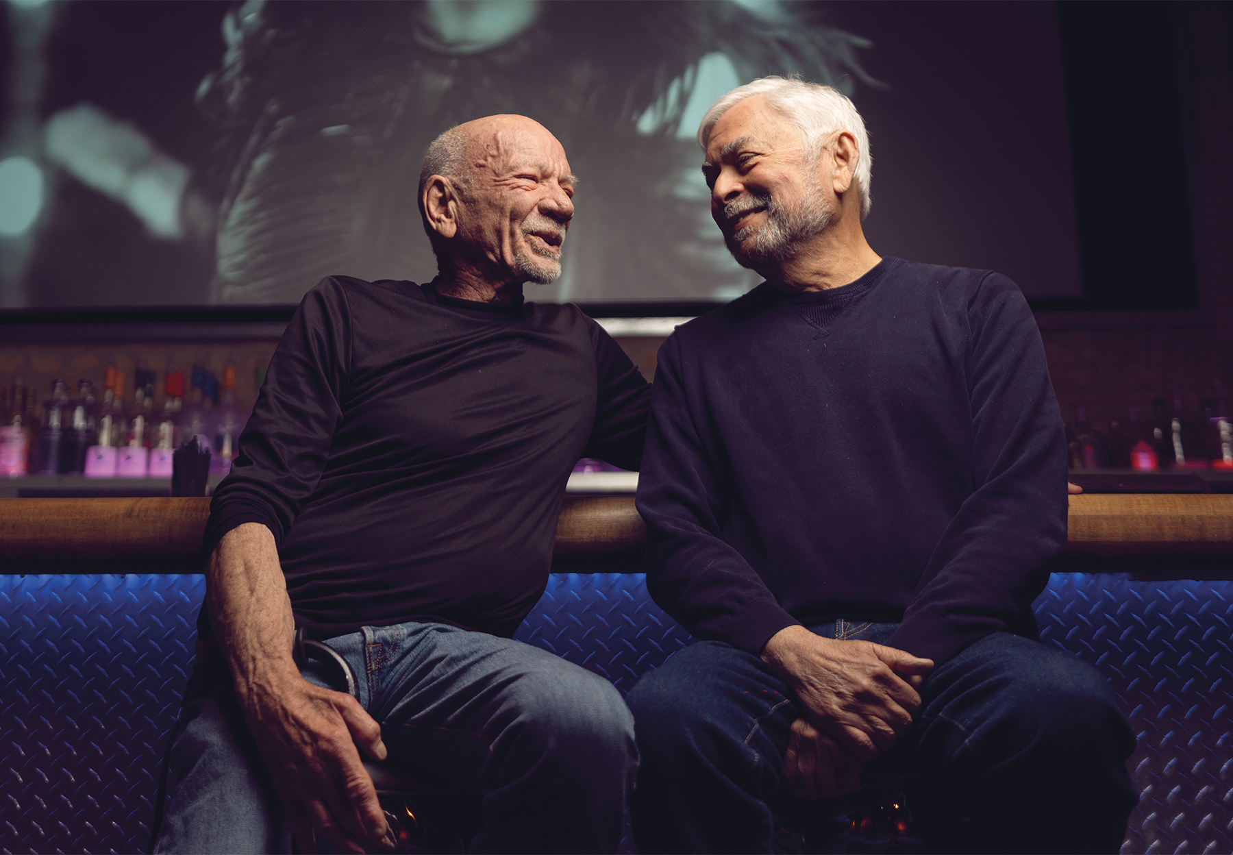 Art Johnston and Pep Peña sit next to each other on bar stools in Sidetrack. They are looking at each other and smiling. Behind them is a large projector screen showing a music video.