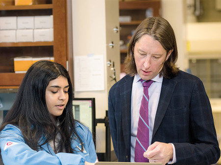 In a lab environment, Julius Lucks, wearing a dark suit and a purple striped tie, stands beside a Northwestern researcher wearing a light blue lab coat. Their mouths are open as though in conversation, and they are gesturing while looking at a laptop screen.