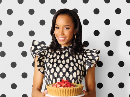 Maya-Camille Broussard smiles at the camera while holding a strawberry basil key lime pie topped with fresh strawberries, on a serving tray. She wears a black and white polka-dotted dress and stands in front of a white and black polka-dotted wall.