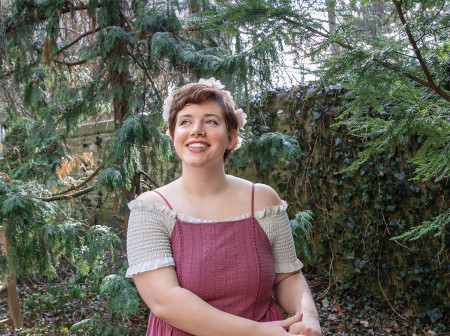 Elizabeth Dudley smiles in a forested area wearing a floral crown and an off-shoulder smocked white top with a dark pink spaghetti-strapped dress over it.