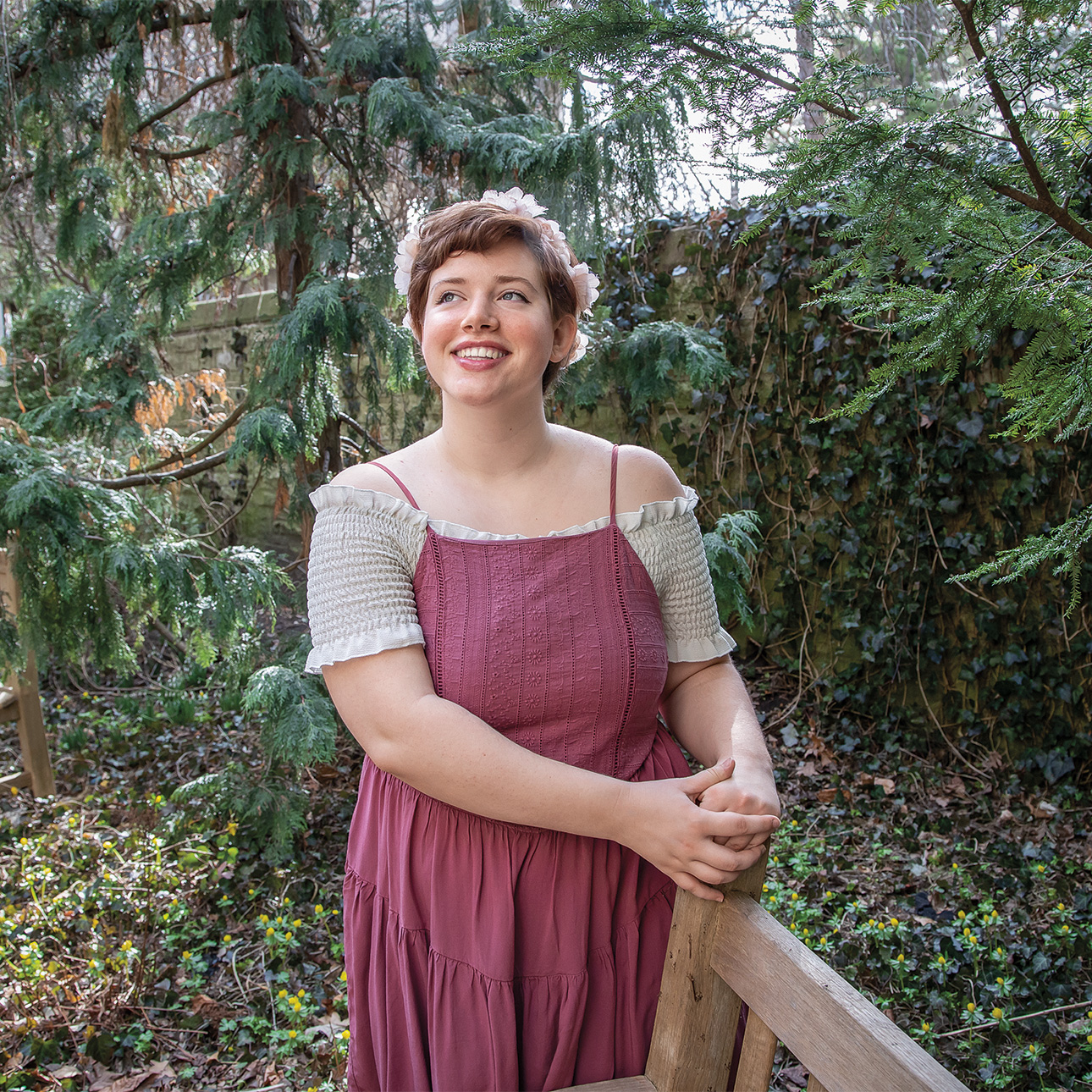 Elizabeth Dudley smiles in a forested area wearing a floral crown and an off-shoulder smocked white top with a dark pink spaghetti-strapped dress over it.