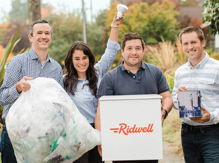 Ridwell founders Ryan Metzger, Aliya Marder, Justin Gough and David Dawson stand outside holding a variety of recyclable items and a Ridwell recycling container.