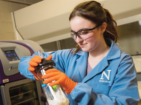 Sarah Sobol, wearing safety glasses, a light blue lab coat with a Northwestern research logo and orange gloves, is conducting synthetic biology research in a lab.