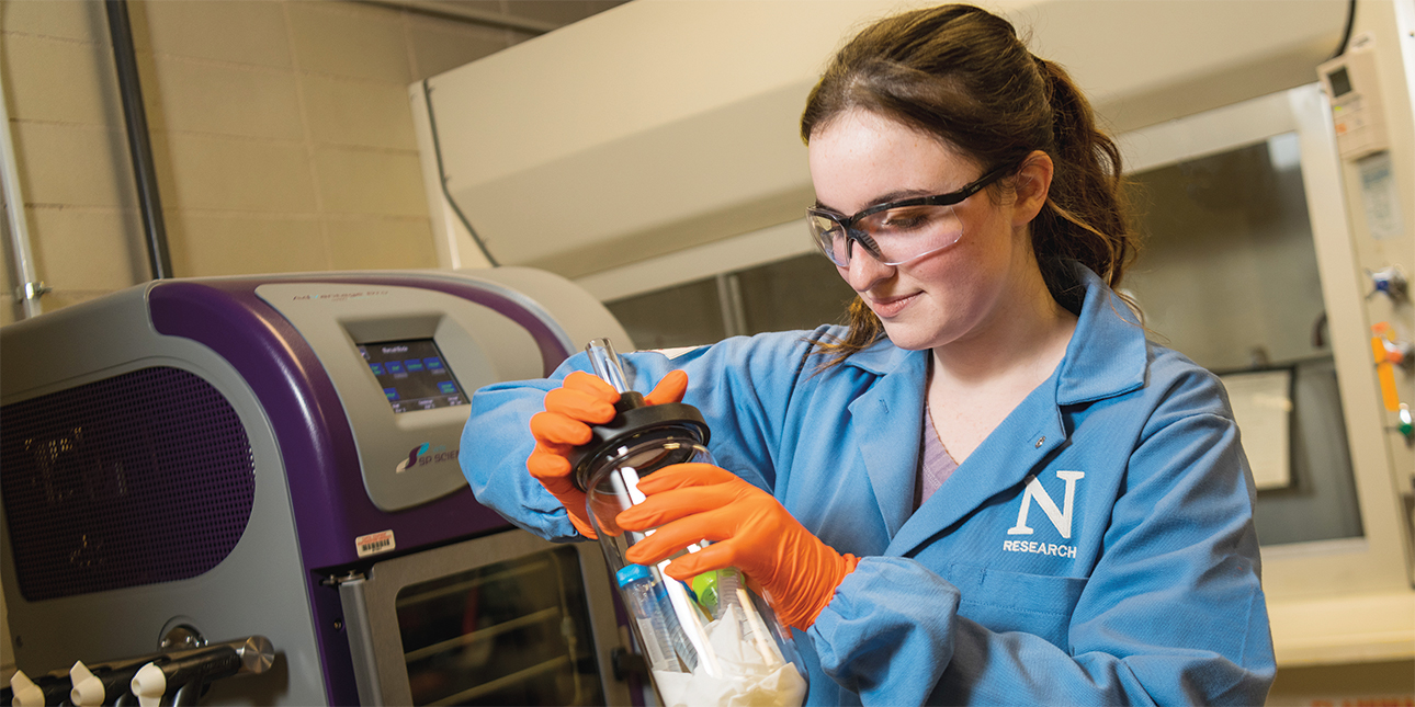 Sarah Sobol, wearing safety glasses, a light blue lab coat with a Northwestern research logo and orange gloves, is conducting synthetic biology research in a lab.