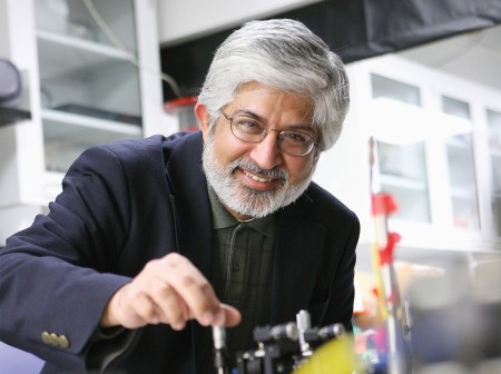 Prem Kumar smiles while standing over a lab bench with his right hand twisting a piece of metal equipment. He wears a blue blazer and a dark green shirt.