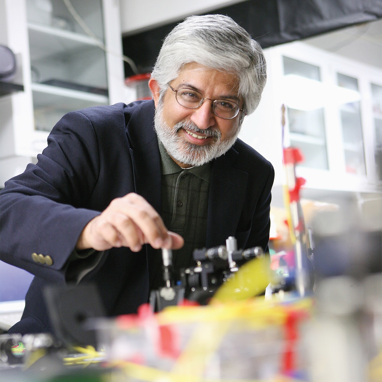 Prem Kumar smiles while standing over a lab bench with his right hand twisting a piece of metal equipment. He wears a blue blazer and a dark green shirt.