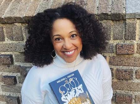 Michelle Foster leans against a brick wall, wearing a white turtleneck top, oblong hoop earrings and a necklace. She is smiling and holding a copy of her book.