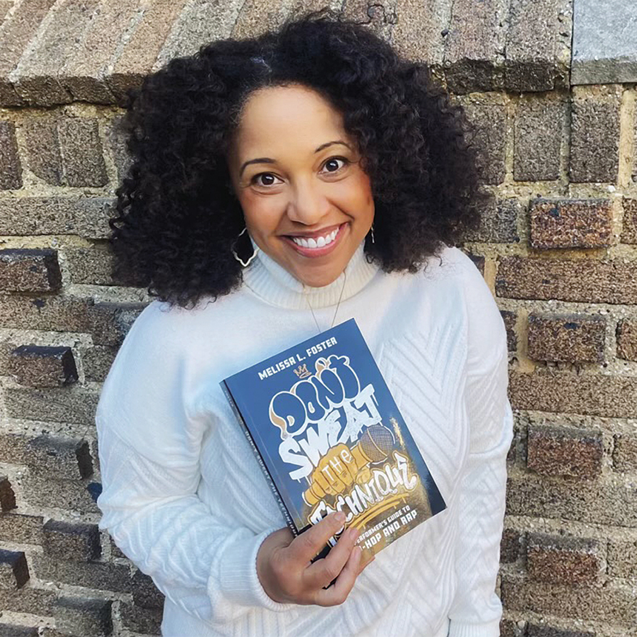 Michelle Foster leans against a brick wall, wearing a white turtleneck top, oblong hoop earrings and a necklace. She is smiling and holding a copy of her book.