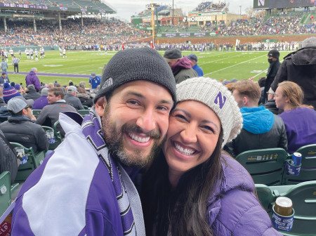 NU Loyal members David Leuchter and Dulce Vasquez pose together in the stands at a Northwestern football game at Wrigley Field, smiling for the camera. Leuchter is wearing a Northwestern jacket and scarf, and Vasquez is wearing a hat with a Northwestern N. A large crowd is visible in the background as well as several football players on the field.