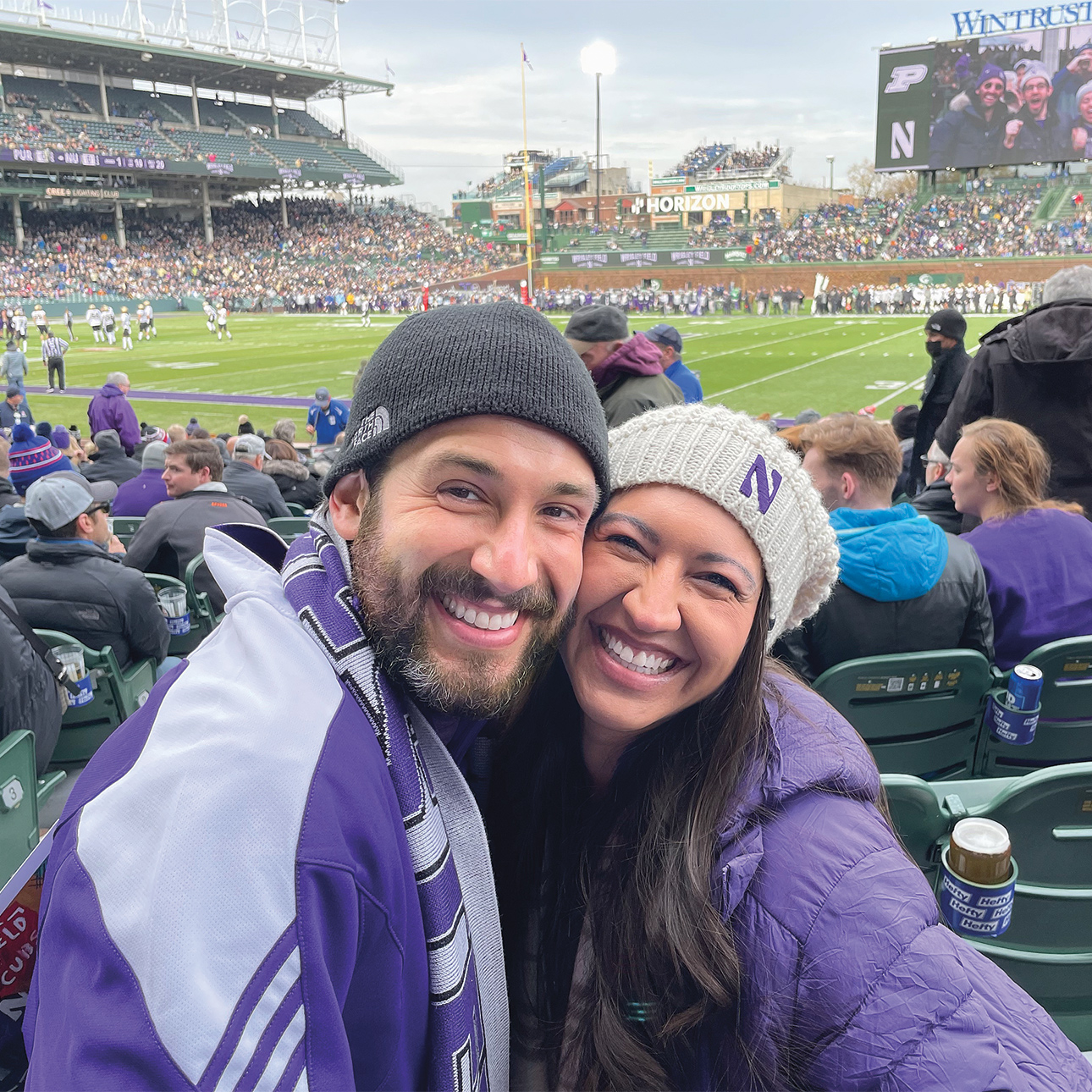NU Loyal members David Leuchter and Dulce Vasquez pose together in the stands at a Northwestern football game at Wrigley Field, smiling for the camera. Leuchter is wearing a Northwestern jacket and scarf, and Vasquez is wearing a hat with a Northwestern N. A large crowd is visible in the background as well as several football players on the field.