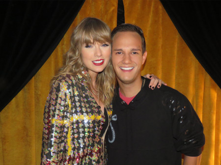 Taylor Swift and Bryan West stand next to each other against a backdrop of golden curtains. Swift’s hand is resting on West’s shoulder. They are both smiling at the camera.