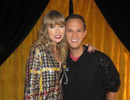 Taylor Swift and Bryan West stand next to each other against a backdrop of golden curtains. Swift’s hand is resting on West’s shoulder. They are both smiling at the camera.