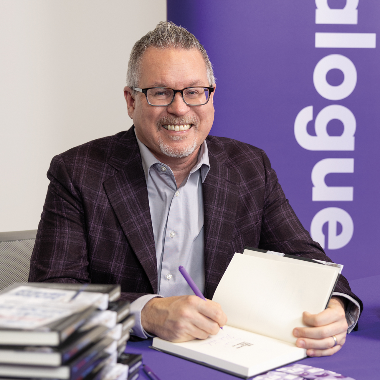 A man wearing glasses and a suit smiles while signing a book.