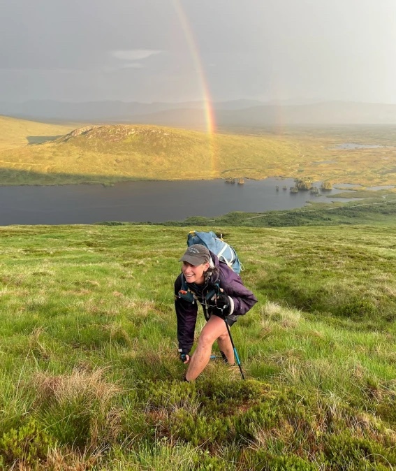Jamie Aarons strides along a grassy stretch of mountain with a body of water behind her and a rainbow stretching into the sky. Here, she is scaling Beinn na Lap in Scotland’s Grampian Mountains, part of the Munros (mountains in Scotland that rise above 3,000 feet)