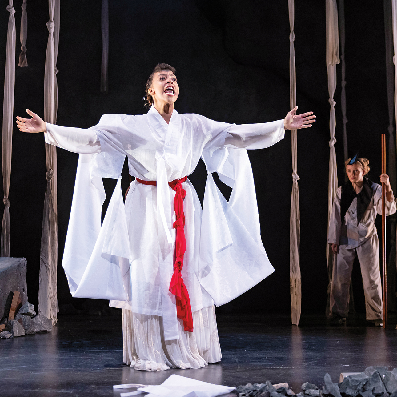 A performer in a white costume with a red belt opens her arms to the audience.