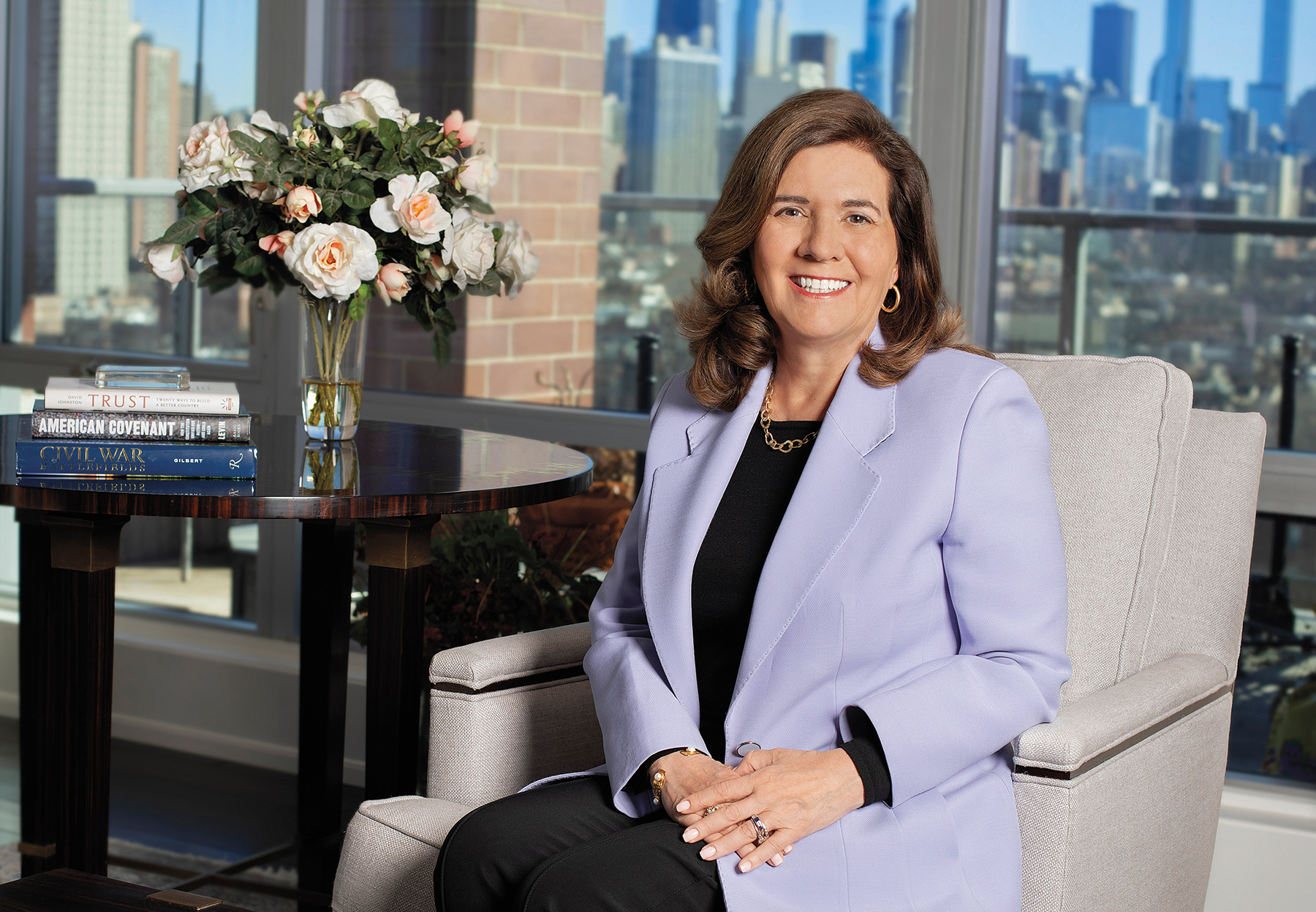 Barbara Stewart smiles at the camera while sitting on a chair next to a sidetable with flowers on it.