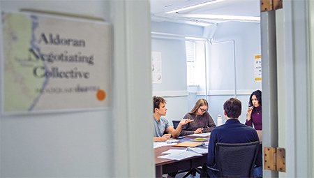 A view into a room from the doorway. Four students sit at a table covered in papers.