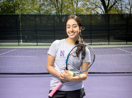 Divya Gupta stands smiling at the camera holding a tennis racquet. She is wearing black shorts and a purple Northwestern shirt.