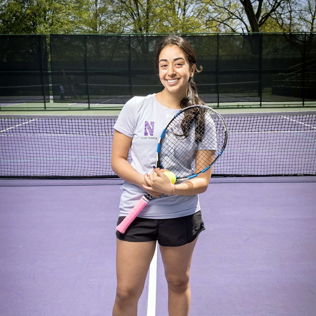Divya Gupta stands smiling at the camera holding a tennis racquet. She is wearing black shorts and a purple Northwestern shirt.
