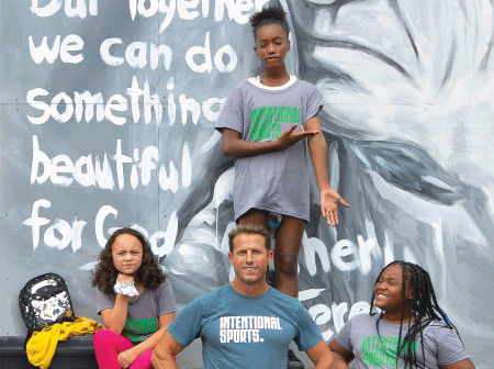 Andy McDermott poses for a photo in front of a mural alongside three kids, all wearing Intentional Sports T-shirts. 