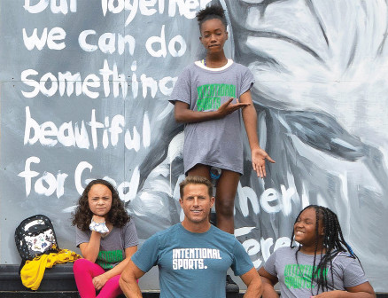 Andy McDermott poses for a photo in front of a mural alongside three kids, all wearing Intentional Sports T-shirts. 