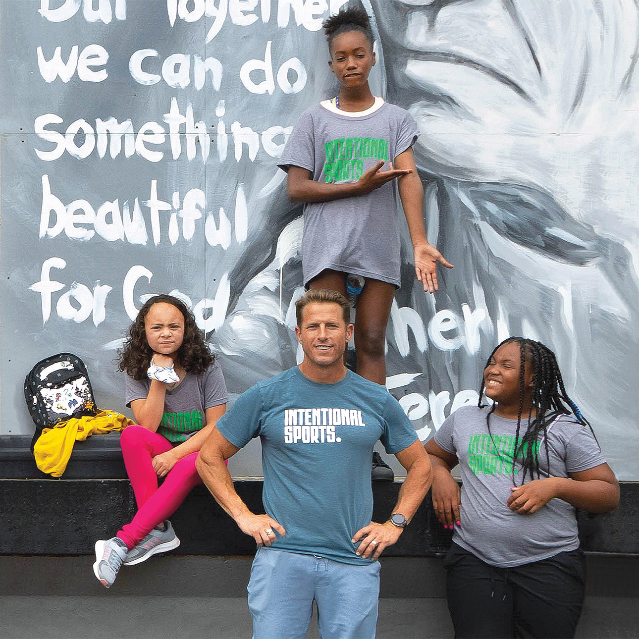 Andy McDermott poses for a photo in front of a mural alongside three kids, all wearing Intentional Sports T-shirts. 