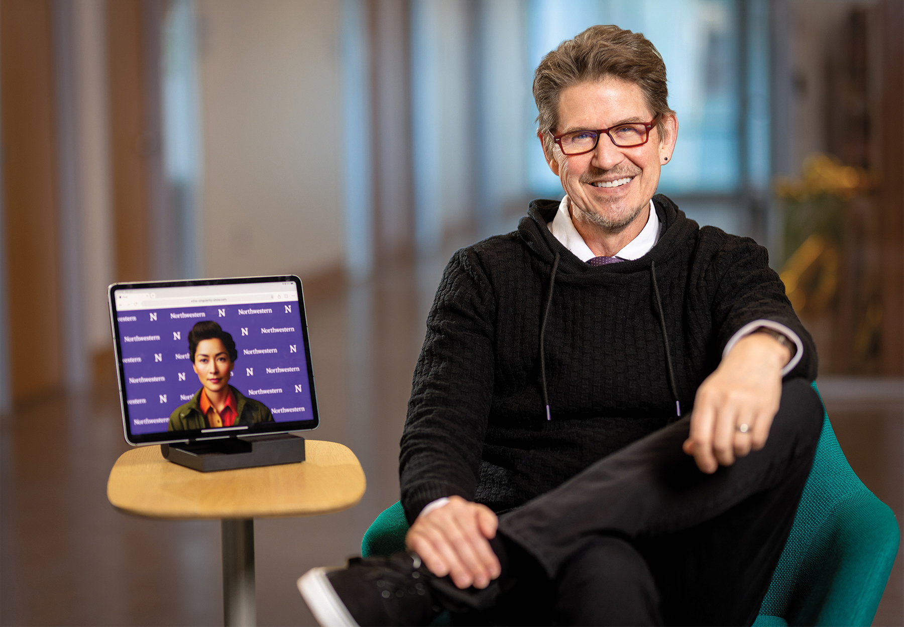 Kris Hammond wears a black hoodie and sits on a green chair. On a table next to him is a tablet showing Elizabeth, a conversational AI avatar.
