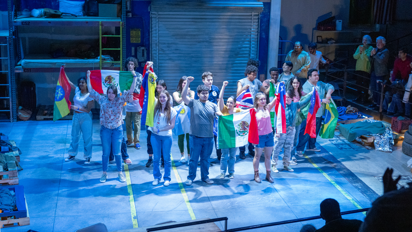 The actors stand together on stage, some raising a fist, some holding flags from different countries. 