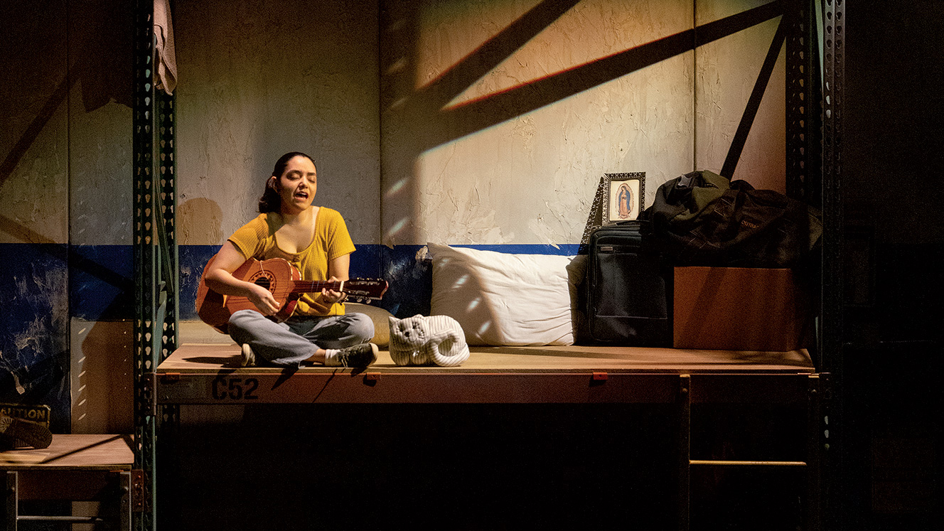 A woman strums a vihuela while sitting on a shelf that has been repurposed as a bunk bed. Her eyes are closed while she sings. 