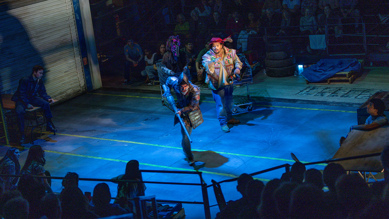 Don Quixote holds a lance on stage and runs forward while Sancho looks on. The audience watches the scene from the seating area in the Barber Theater. 
