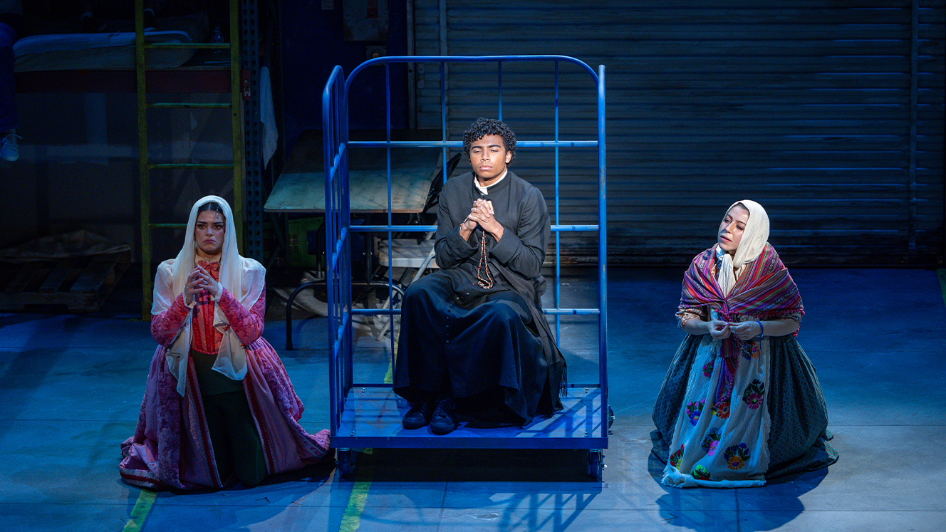Two women in costumes kneel on either side of a dolly, with their hands clasped in prayer. On the dolly, a person dressed as a friar clasps his hands in prayer. 