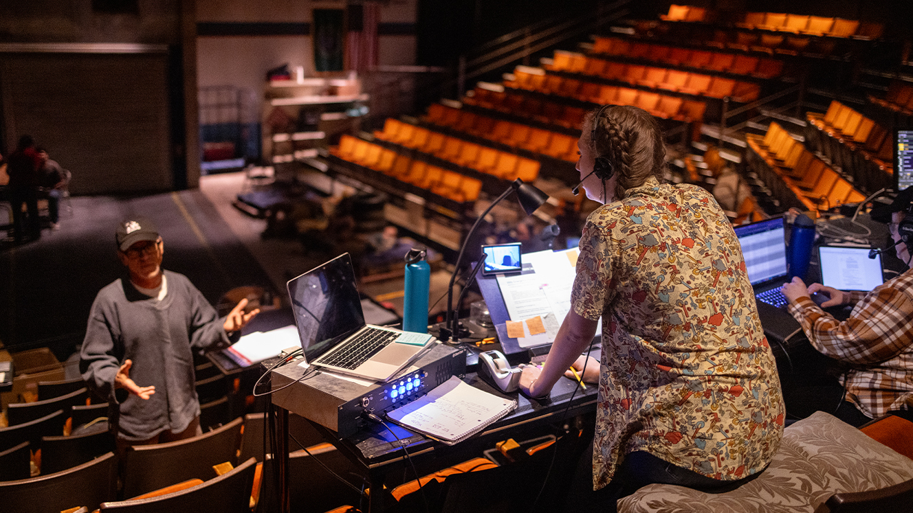 Audrey Michael sits at a table in the audience seating area of the Barber Theater, facing away from the camera. Henry Godinez is a few rows below her, gesturing with his hands.